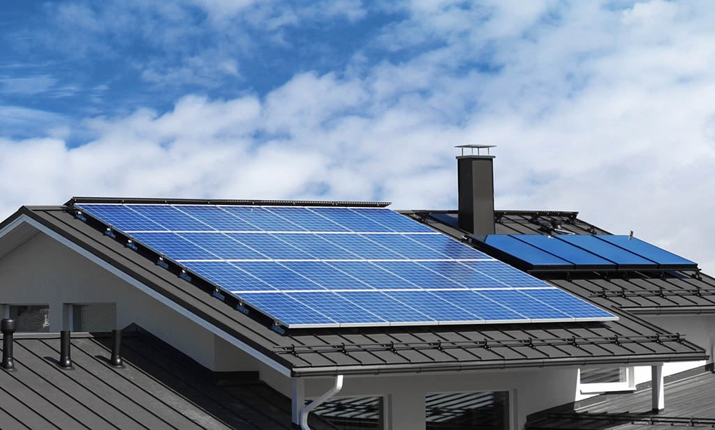 Instalacja paneli słonecznych na ciemnym, spadzistym dachu domu jednorodzinnego z kominem, na tle błękitnego nieba z chmurami.