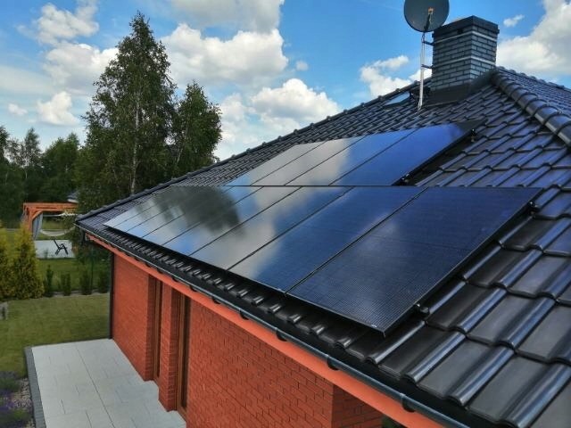 Czarny dach z dachówki ceramicznej z zamontowanymi panelami słonecznymi, widoczny komin, antena satelitarna oraz fragment elewacji z czerwonej cegły i trawnik