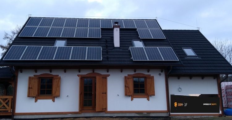 Dom jednorodzinny z ciemną dachówką i panelami słonecznymi na dachu, okna z drewnianymi okiennicami, częściowo odsłonięta brama garażowa z logo firmy.