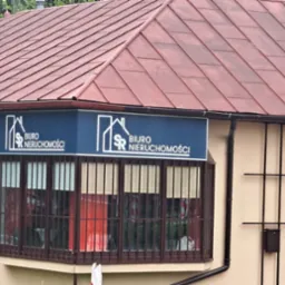 Budynek biura nieruchomości z brązowym dachem i niebieskim szyldem z białym logo i napisem 'Biuro Nieruchomości'. Okna z kratami. Na drzwiach informacja o usługach i kontakt.