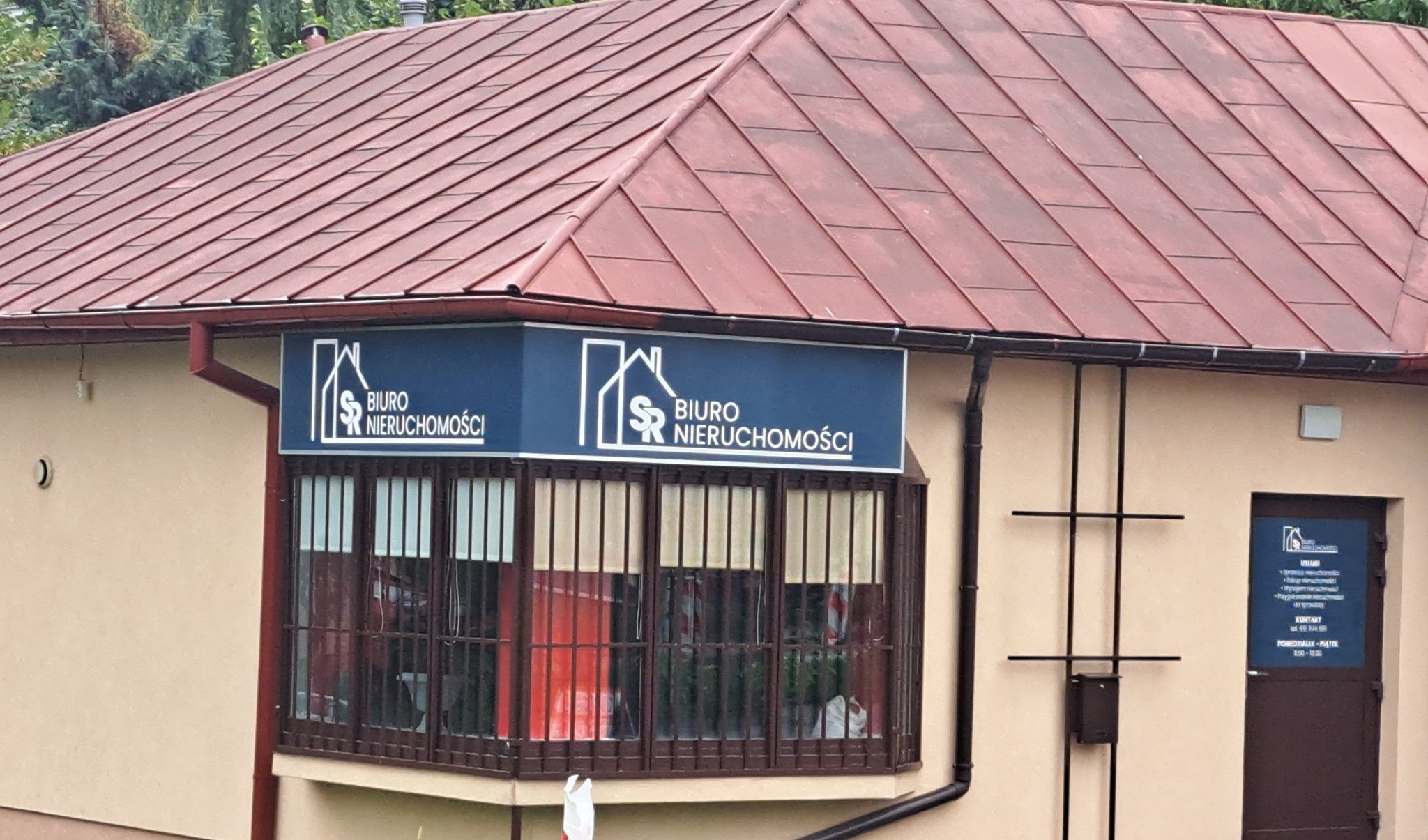 Budynek biura nieruchomości z brązowym dachem i niebieskim szyldem z białym logo i napisem 'Biuro Nieruchomości'. Okna z kratami. Na drzwiach informacja o usługach i kontakt.