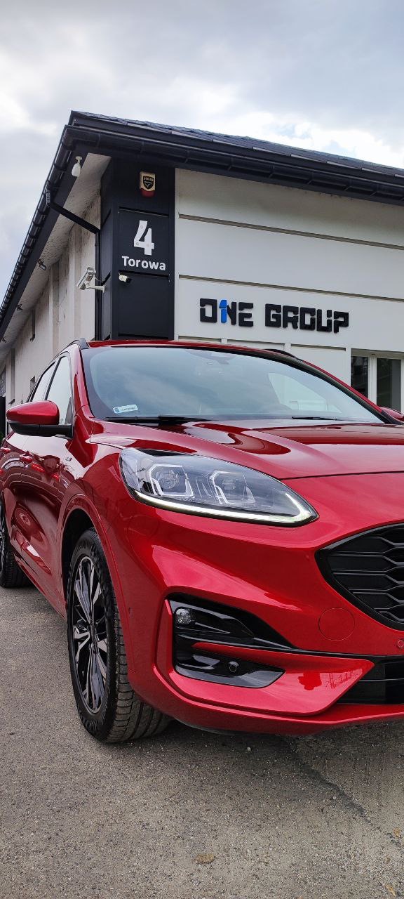 Czerwony samochód Ford Kuga zaparkowany przed budynkiem firmy ONE GROUP z adresem Torowa 4, widok z przodu, lekko z boku.