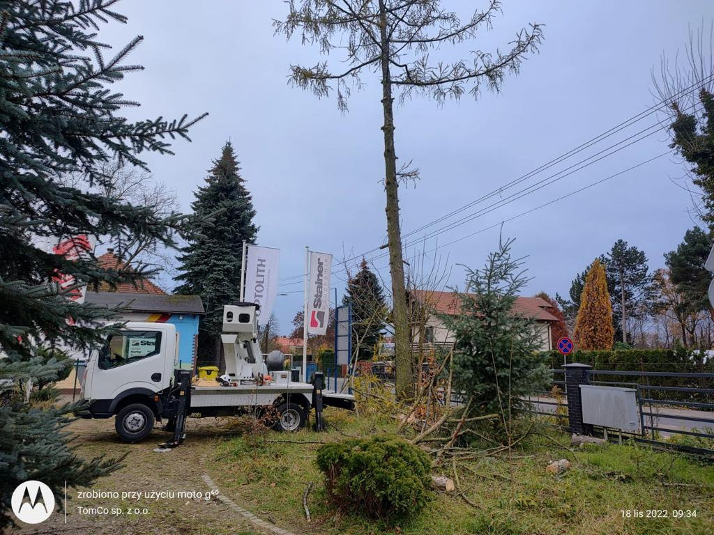 Specjalistyczny pojazd z podnośnikiem hydraulicznym na posesji po wycince drzewa, widoczne ścięte gałęzie, w tle budynki mieszkalne i banery reklamowe firm.