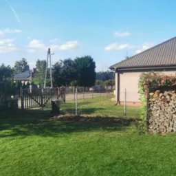 Widok na zadbany ogr&oacute;d z trawnikiem, drewnianą bramką i drewutnią obrośniętą bluszczem, pełną ułożonego drewna opałowego, z domem w tle.