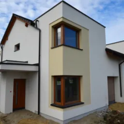 Nowoczesny, szeregowy dom murowany z jasną elewacją, drewnianymi oknami i elementami wykończeniowymi, z widocznymi przewodami elektrycznymi na ziemi.