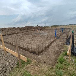 Szerbud - Przygotowanie terenu pod fundamenty: wykopane rowy, wbite pręty zbrojeniowe, drewniane szalunki, robotnik podlewa ziemię w słoneczny dzień.