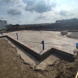 Szerbud - Wylany fundament pod budynek jednorodzinny z widocznymi pionowymi rurami instalacyjnymi i zbrojeniami, na tle częściowo zagospodarowanego terenu budowy i domu w stanie surowym.