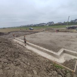 Szerbud - Przygotowanie fundamentów pod budowę, widoczny zarys betonowych ścian fundamentowych, prace ziemne w toku, koparka i pracownicy na placu budowy, w tle zabudowa jednorodzinna.