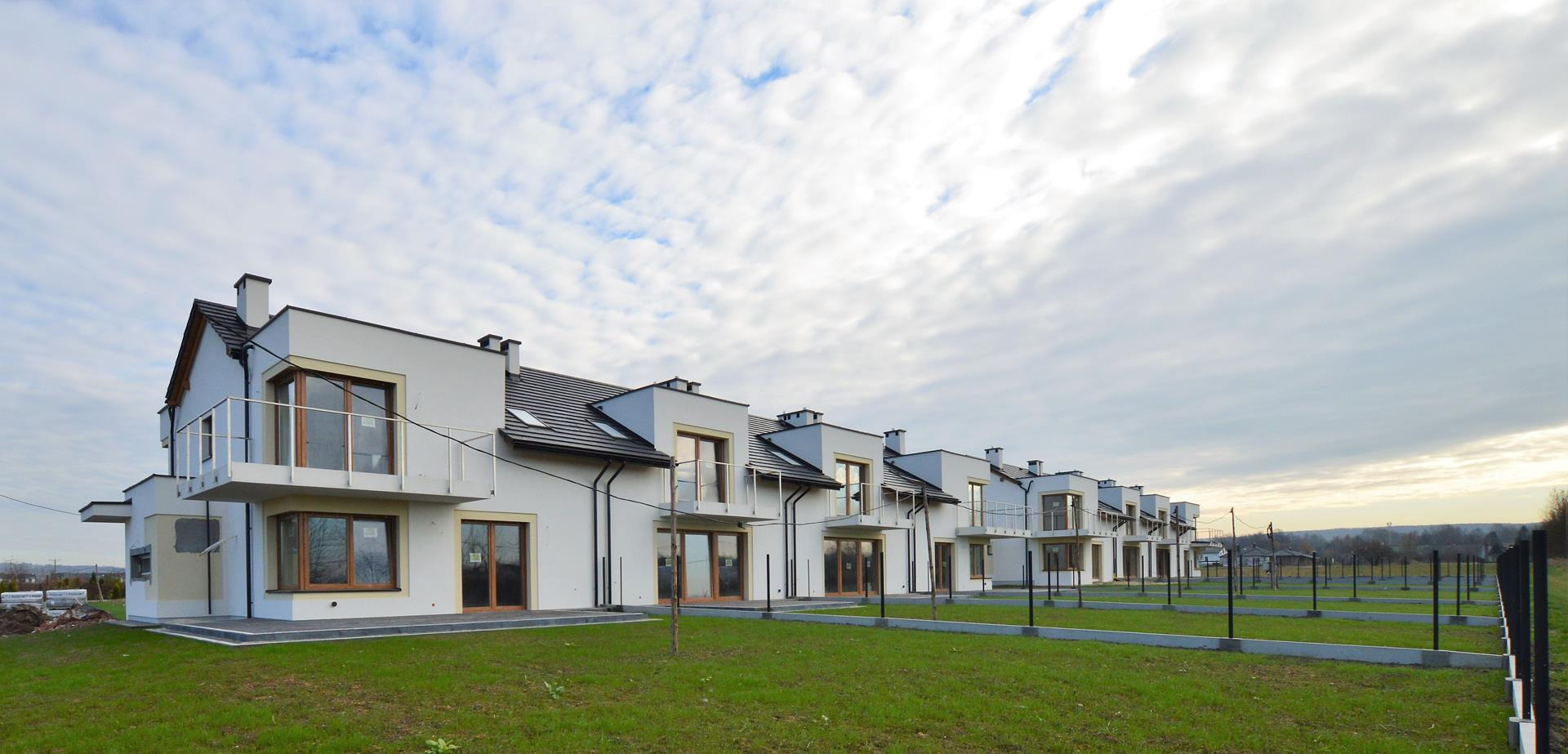 Szereg identycznych, nowoczesnych, dwukondygnacyjnych budynków z balkonami, wykończonych na biało, z ciemnymi dachami i drewnianymi ramami okiennymi, na trawniku z elementami ogrodzenia.