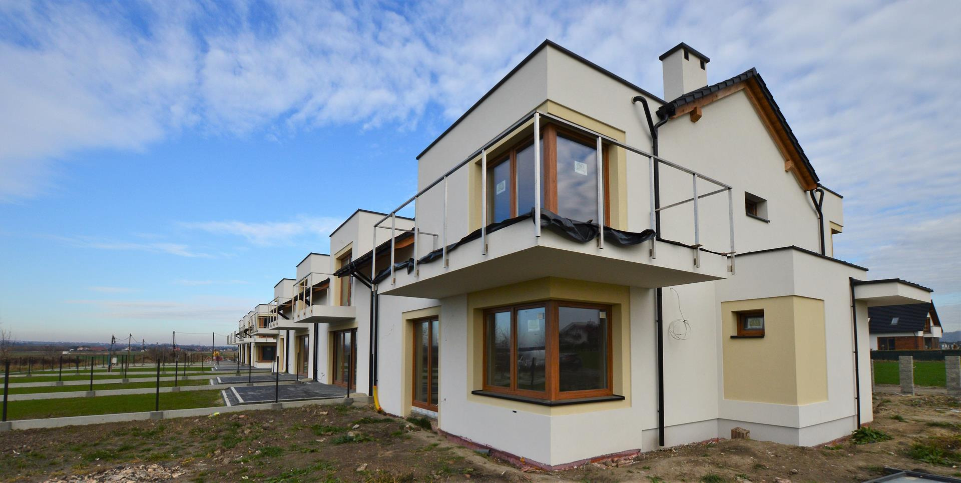 Szeregówka nowych, murowanych domów jednorodzinnych z balkonami, w stanie deweloperskim, na tle błękitnego nieba z chmurami.