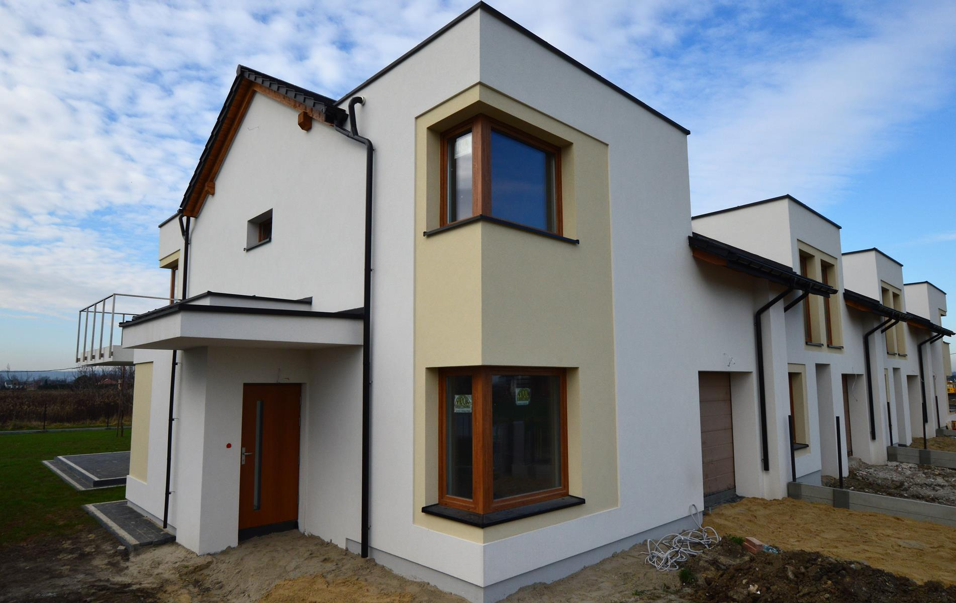 Nowoczesny, szeregowy dom murowany z jasną elewacją, drewnianymi oknami i elementami wykończeniowymi, z widocznymi przewodami elektrycznymi na ziemi.