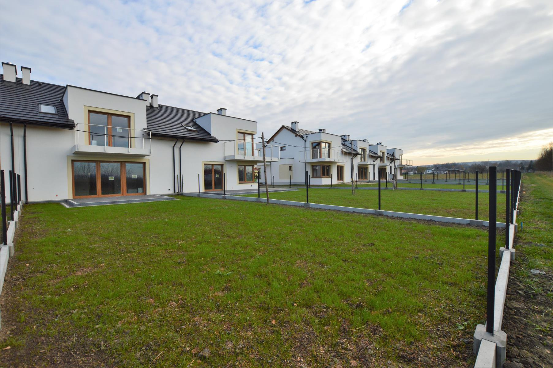 Szereg nowo wybudowanych, białych domów z balkonami i ciemnymi dachami, oddzielonych czarnym ogrodzeniem, na tle pochmurnego nieba.