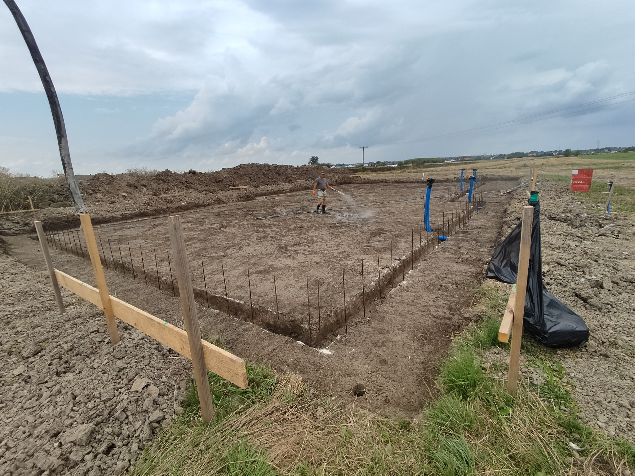 Przygotowanie terenu pod fundamenty: wykopane rowy, wbite pręty zbrojeniowe, drewniane szalunki, robotnik podlewa ziemię w słoneczny dzień.