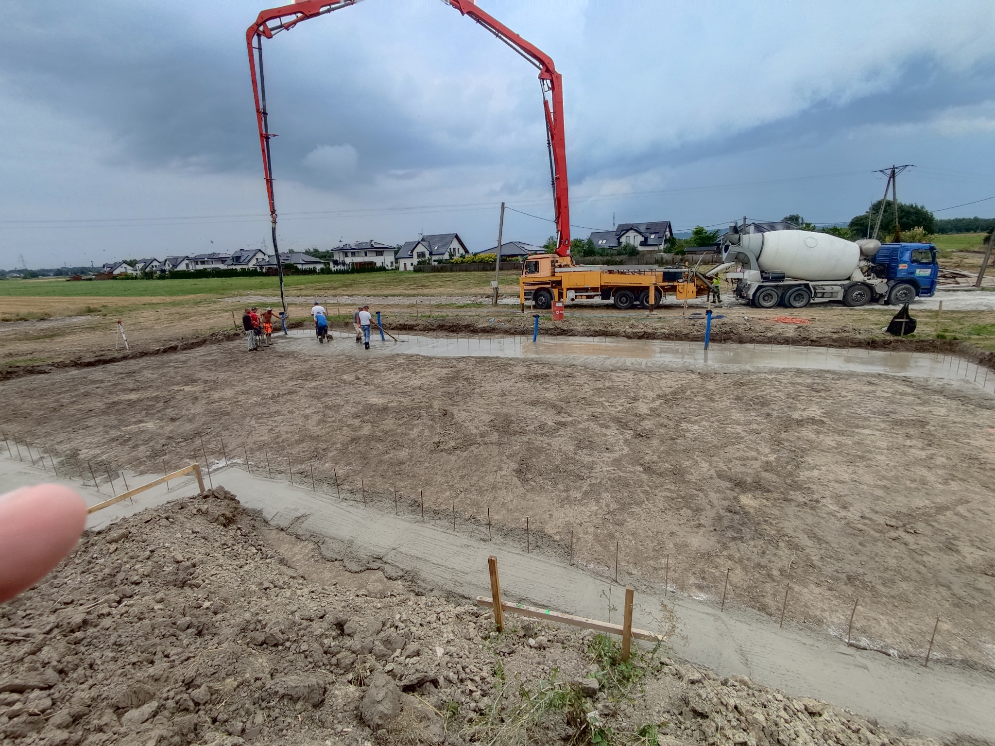 Wylewanie fundamentów pod budowę domu z użyciem pompy do betonu i betoniarki, widoczni pracownicy budowlani.