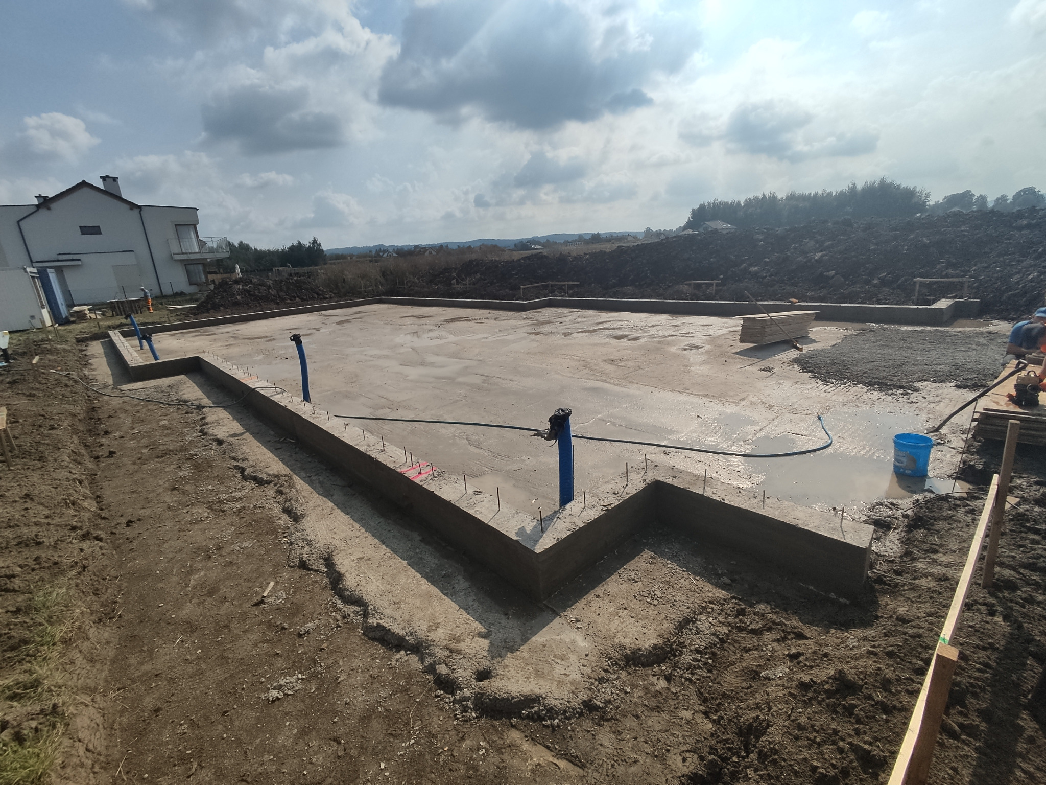 Wylany fundament pod budynek jednorodzinny z widocznymi pionowymi rurami instalacyjnymi i zbrojeniami, na tle częściowo zagospodarowanego terenu budowy i domu w stanie surowym.