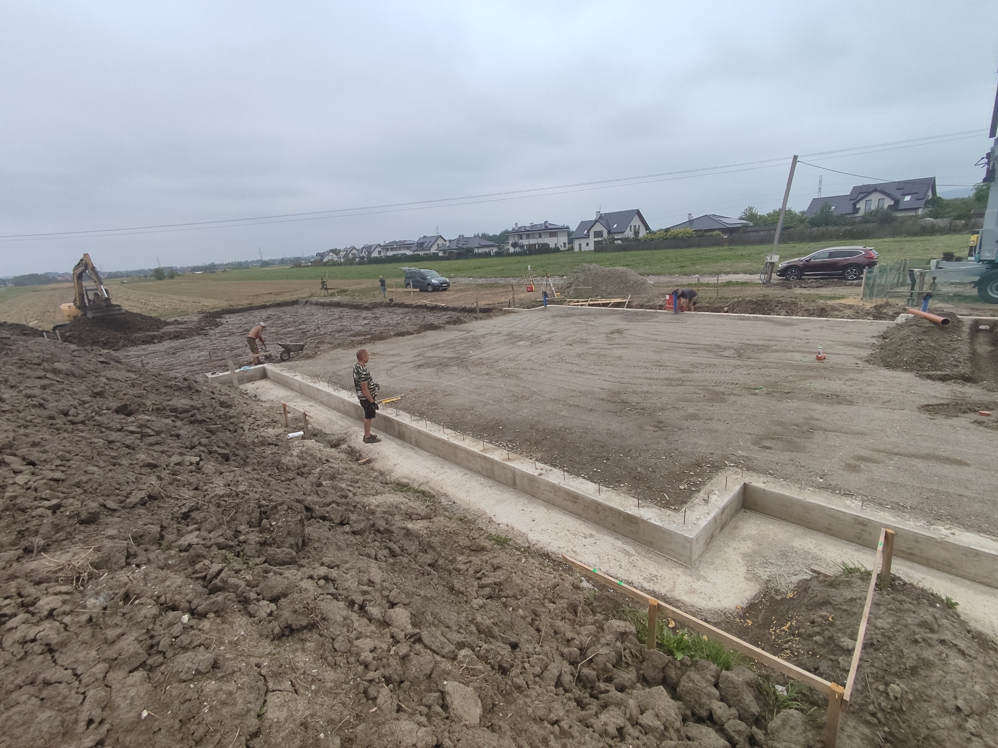 Przygotowanie fundamentów pod budowę, widoczny zarys betonowych ścian fundamentowych, prace ziemne w toku, koparka i pracownicy na placu budowy, w tle zabudowa jednorodzinna.