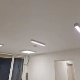 Białe, prostokątne lampy LED zamontowane na suficie w pomieszczeniu z oknami i drzwiami. Widoczny czujnik dymu i kable przy oknie. Minimalistyczny design oświetlenia.
