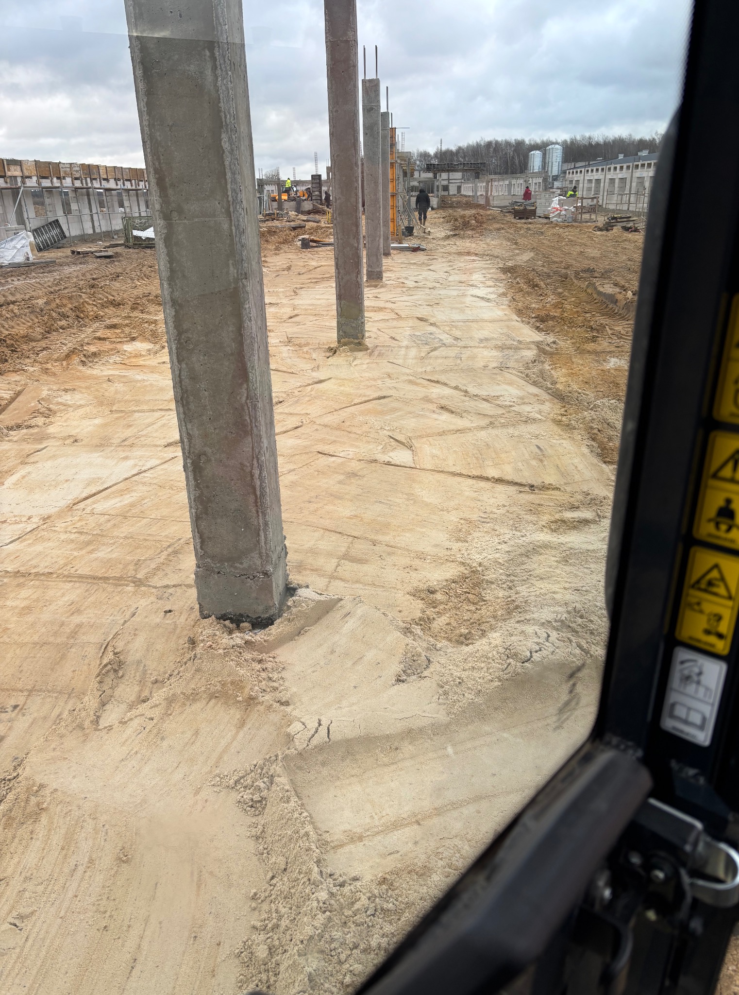 Wykopane fundamenty pod budynek z widocznymi słupami nośnymi, teren przygotowany pod wylewkę. Prace ziemne w toku, widok z kabiny maszyny.