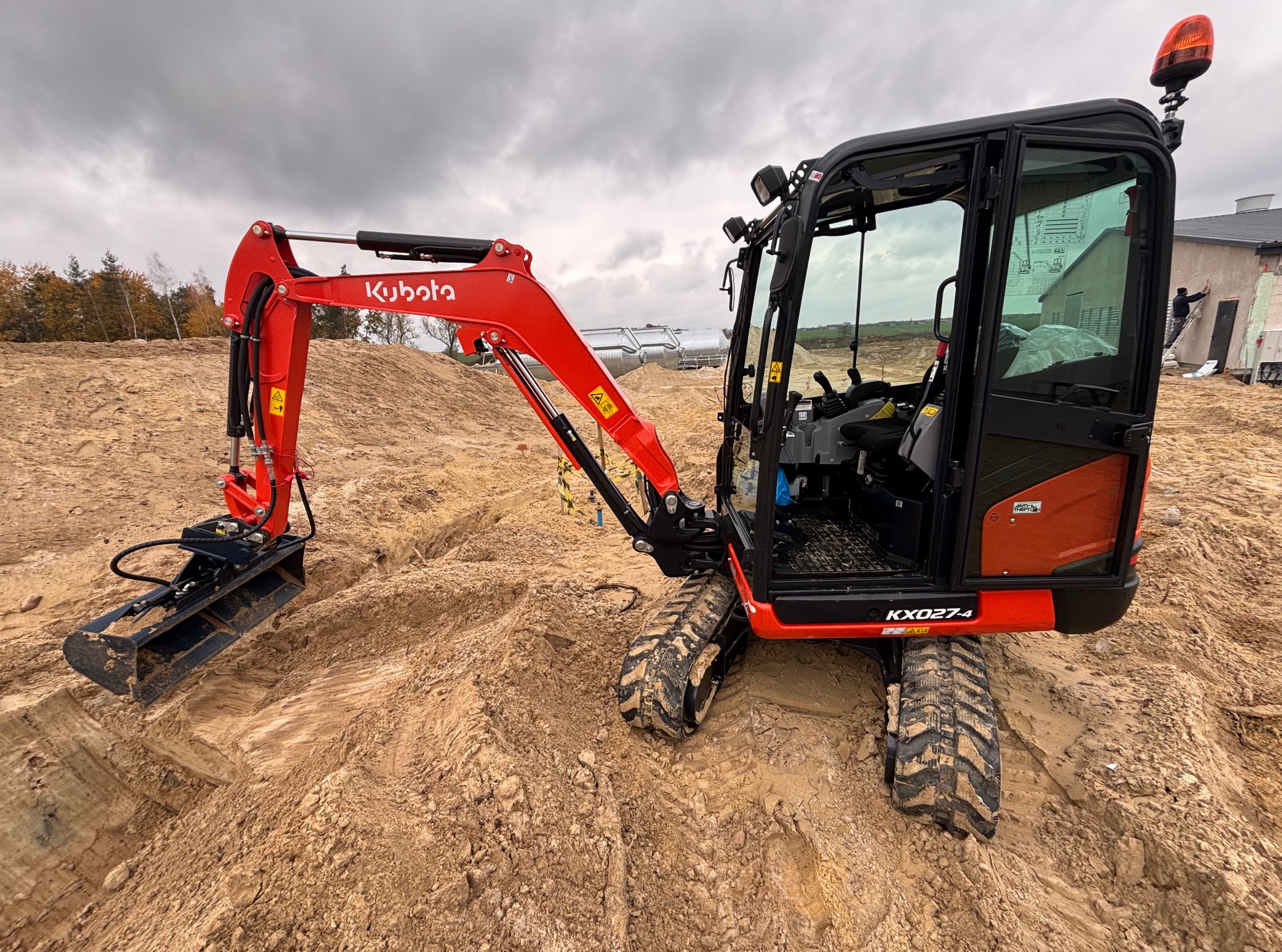 Czerwona minikoparka Kubota KX027-4 na placu budowy z wykopanym rowem, w tle budynek w trakcie prac wykończeniowych. Pochmurne niebo.