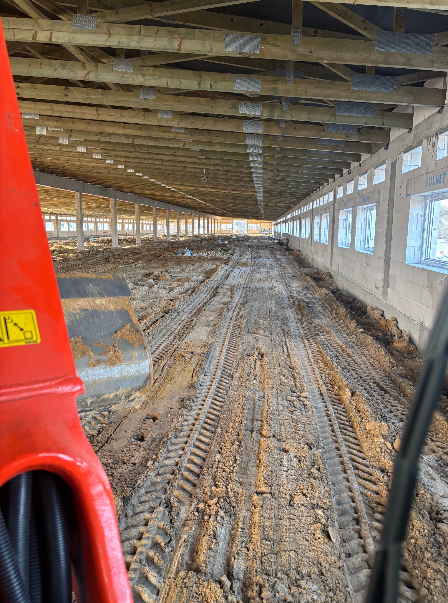 Wnętrze hali w budowie z widocznymi śladami opon na ziemi i fragmentem pomarańczowej maszyny budowlanej w kadrze. Widoczne okna i konstrukcja dachu.