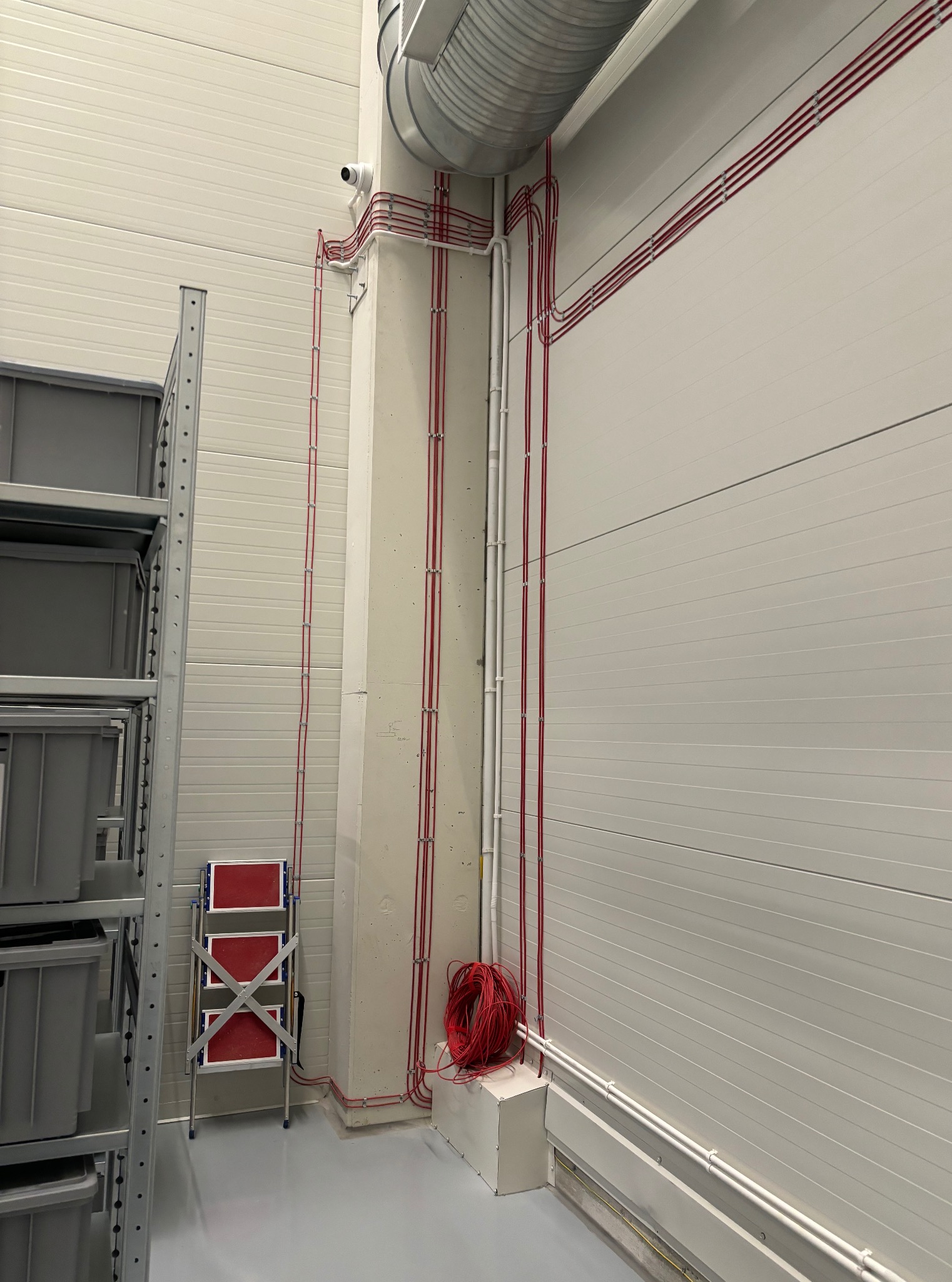 Czerwone przewody elektryczne równolegle ułożone na ścianie w hali, zabezpieczone uchwytami. Przy podłodze zwój kabli i puszka. Metalowy regał i drabina w tle.