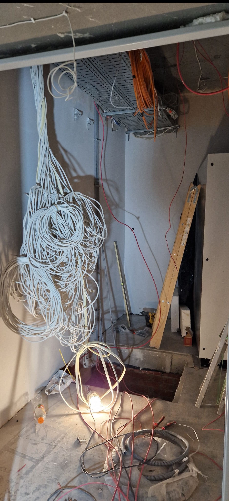 Wnętrze w trakcie prac elektrycznych: zwisające kable, koryta kablowe pod sufitem, drabina oparta o ścianę, narzędzia i lampa oświetlająca podłoże.