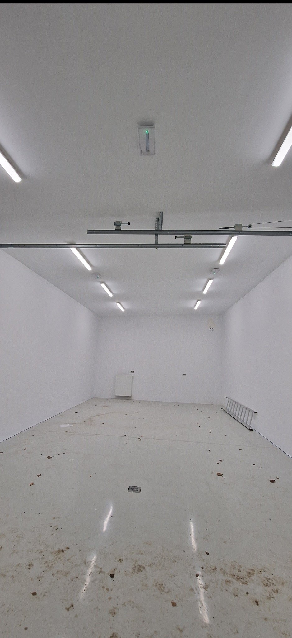 Wnętrze hali z białymi ścianami i podłogą, oświetlone rzędami lamp. Widoczna drabina oparta o ścianę. Podłoga lekko zabrudzona. Minimalistyczny design.