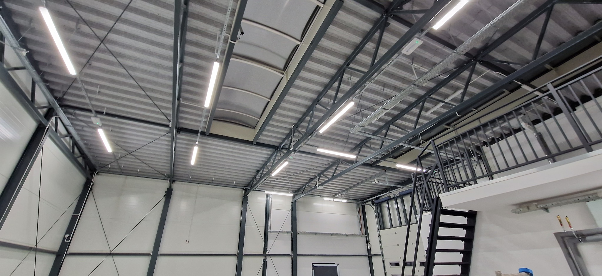 Wnętrze hali przemysłowej z zamontowanym oświetleniem LED. Widoczne metalowe konstrukcje dachu, białe ściany i antresola ze schodami. Górne światło dzienne przez świetliki.