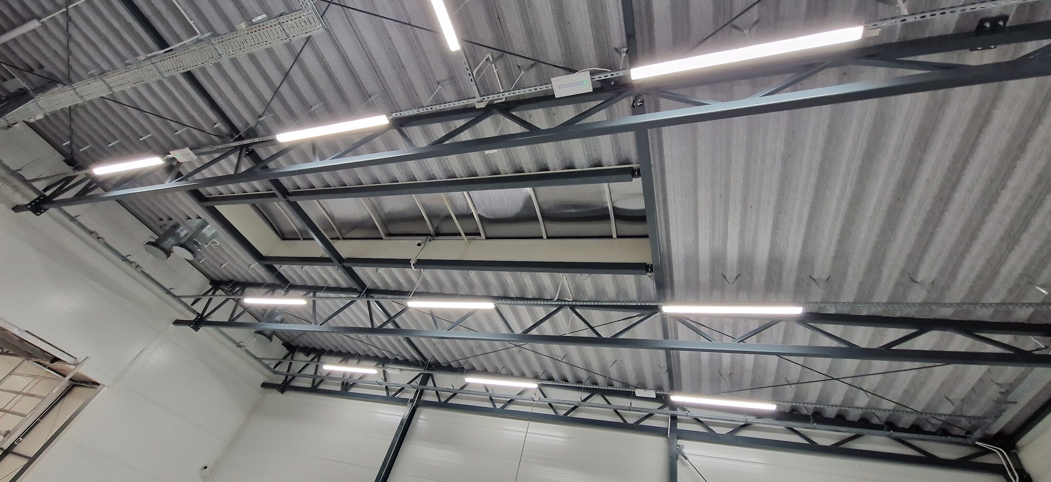 Zamontowane, liniowe oświetlenie LED pod stropem z blachy trapezowej w hali. Widoczne elementy konstrukcji nośnej i okablowanie.