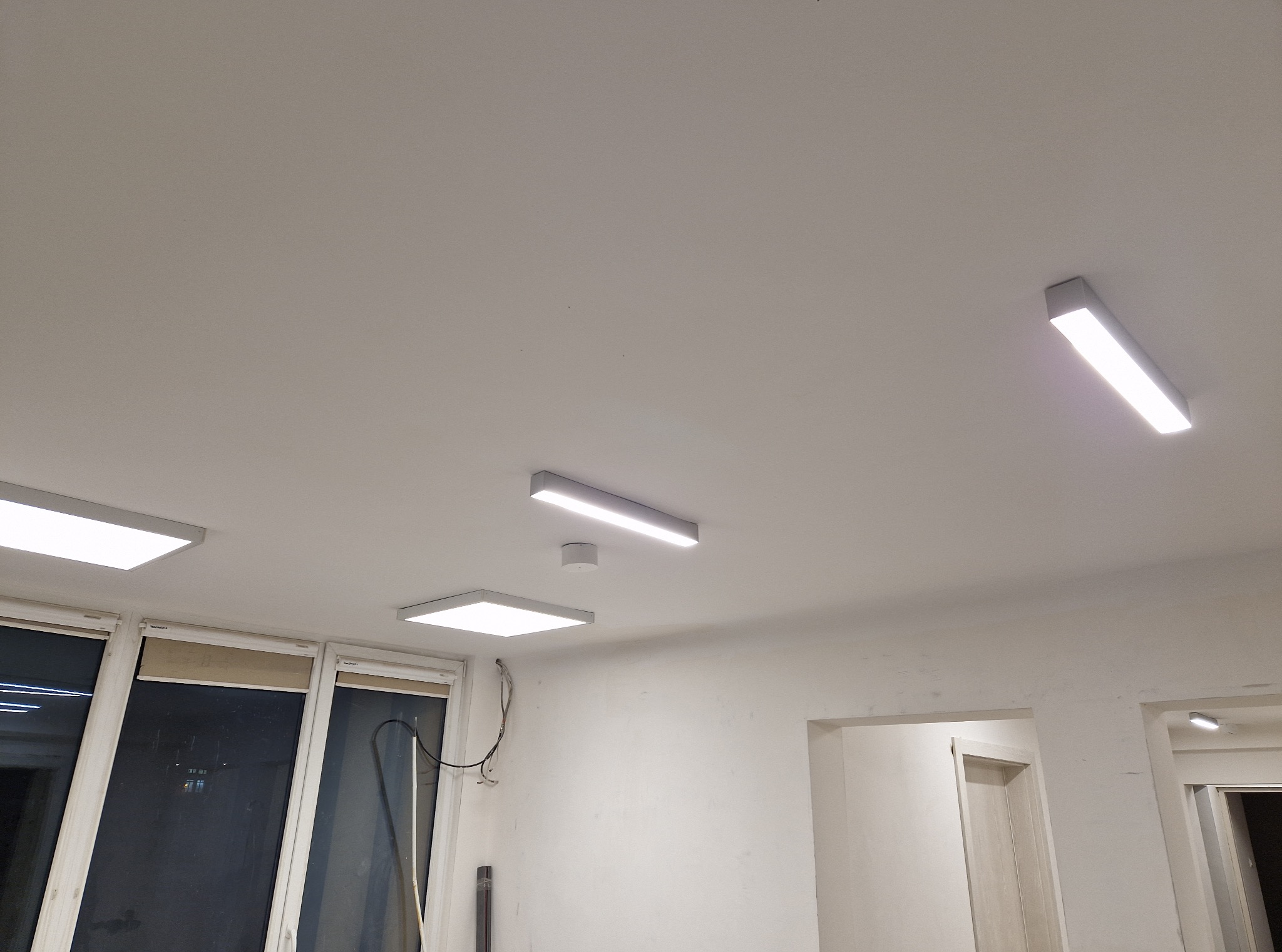 Białe, prostokątne lampy LED zamontowane na suficie w pomieszczeniu z oknami i drzwiami. Widoczny czujnik dymu i kable przy oknie. Minimalistyczny design oświetlenia.