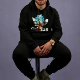 Mężczyzna siedzi na stołku barowym ubrany w czarny dres z kapturem z nadrukiem postaci z anime, złoty zegarek na nadgarstku, szare buty sportowe, na fioletowym tle.