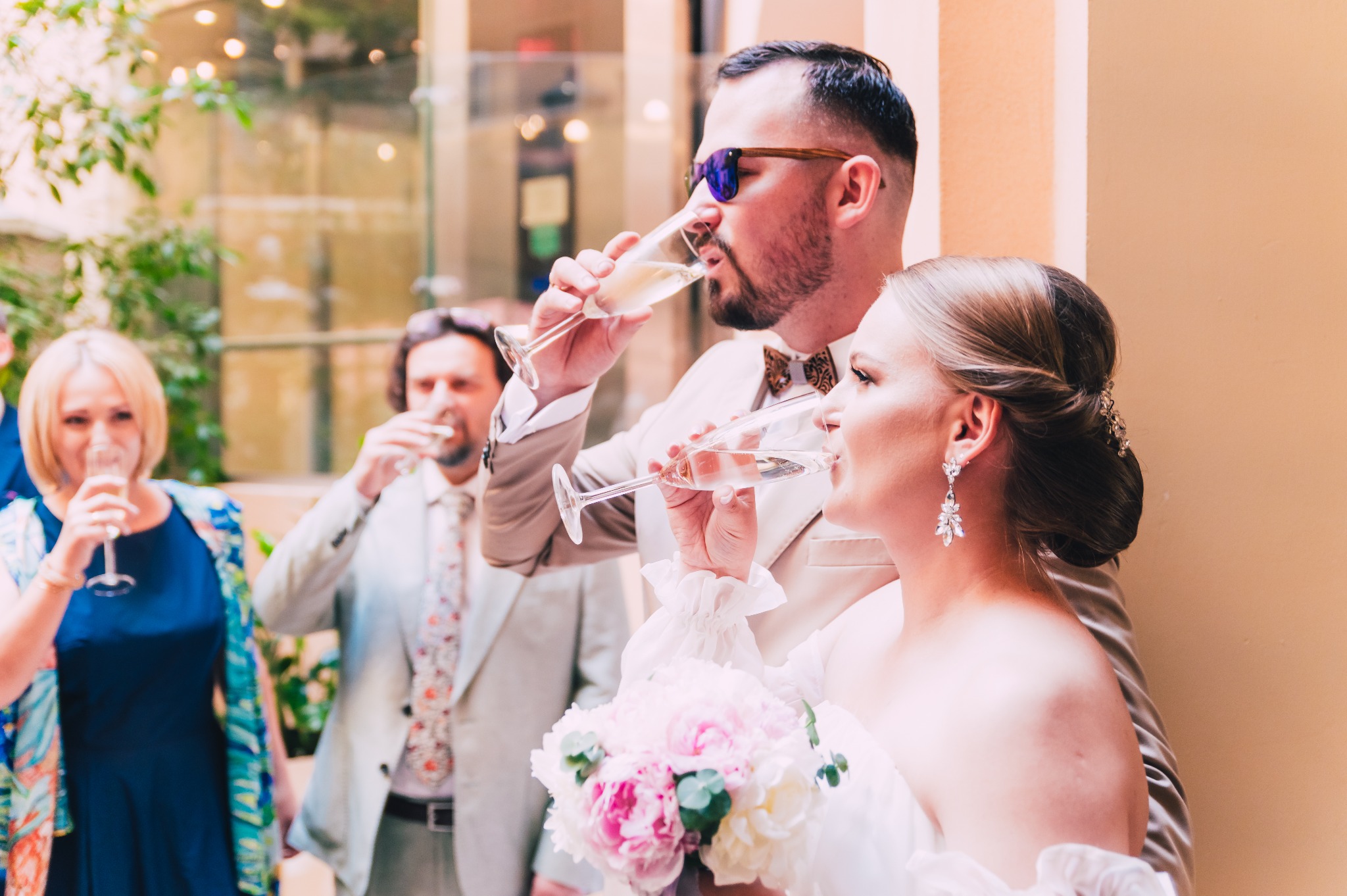 Para młoda w jasnych strojach pije szampana z kieliszków na tle gości weselnych, mężczyzna w okularach przeciwsłonecznych i drewnianej muszce, panna młoda z bukietem piwonii.