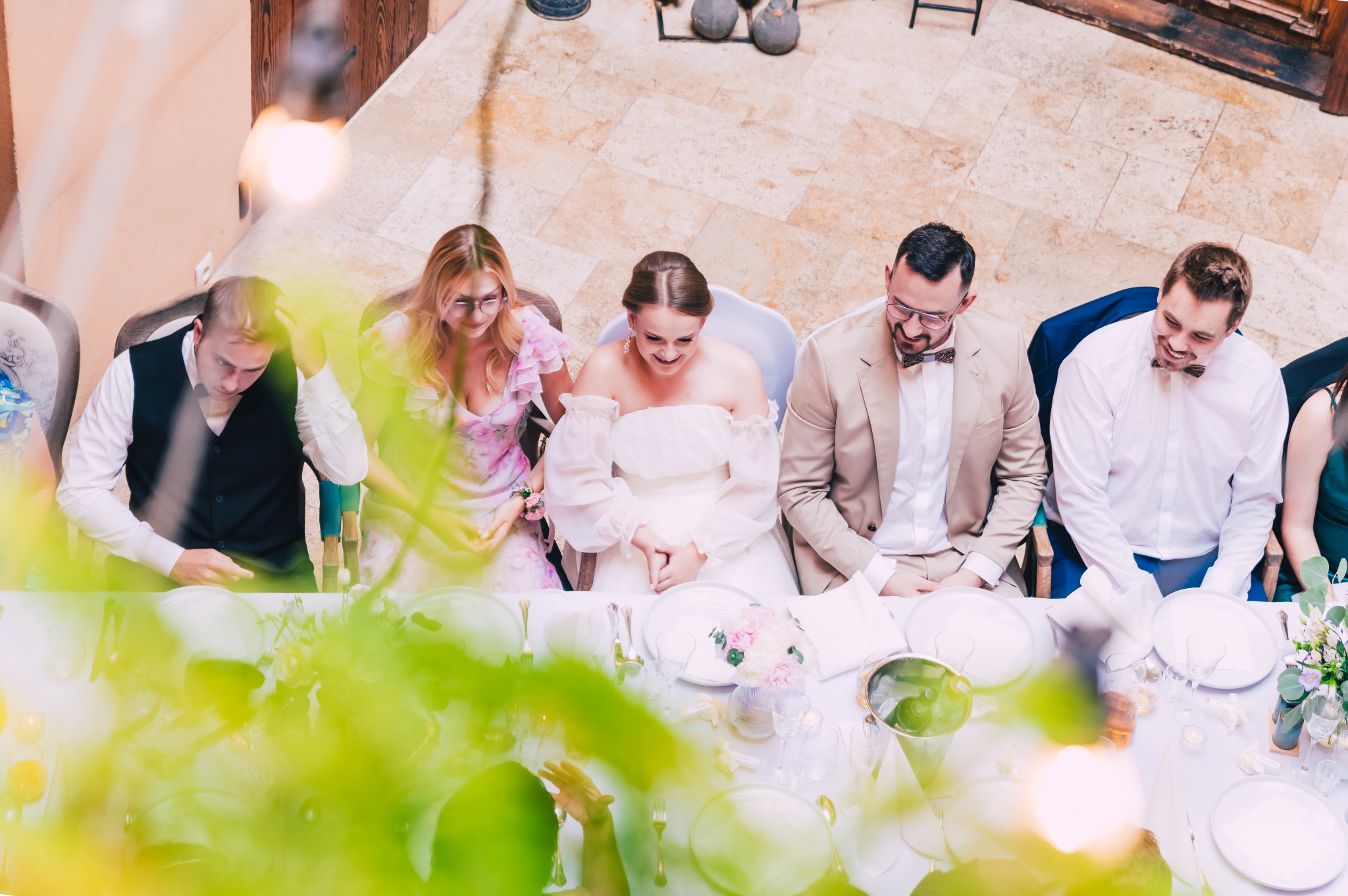 Widok z góry na parę młodą i gości siedzących przy stole weselnym z białym obrusem, kwiatami i nakryciami, w tle kamienna podłoga i drewniane drzwi.