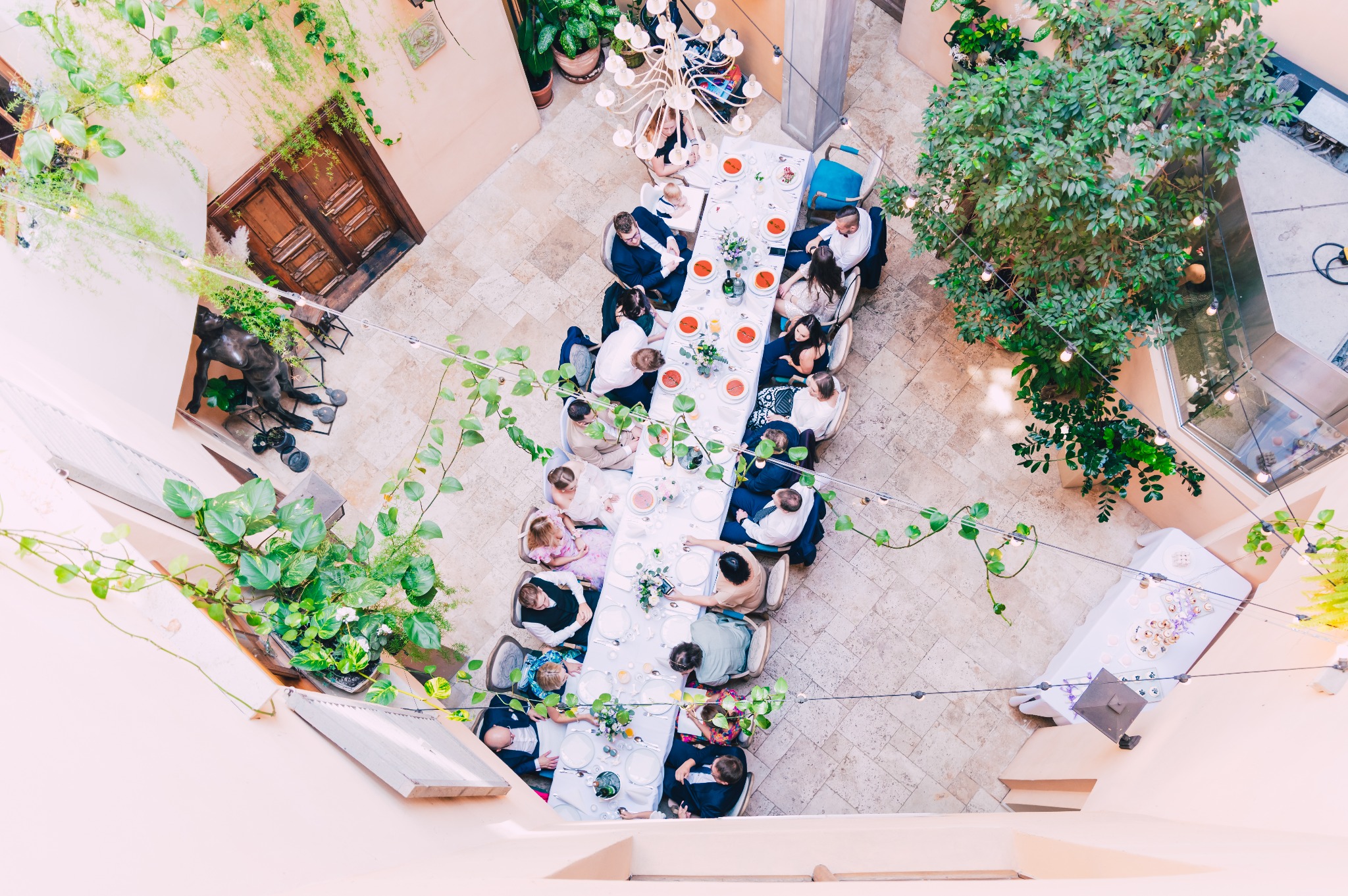 Widok z góry na długi stół z gośćmi w eleganckiej sali z roślinami, rzeźbą i żyrandolem, prawdopodobnie podczas przyjęcia weselnego.