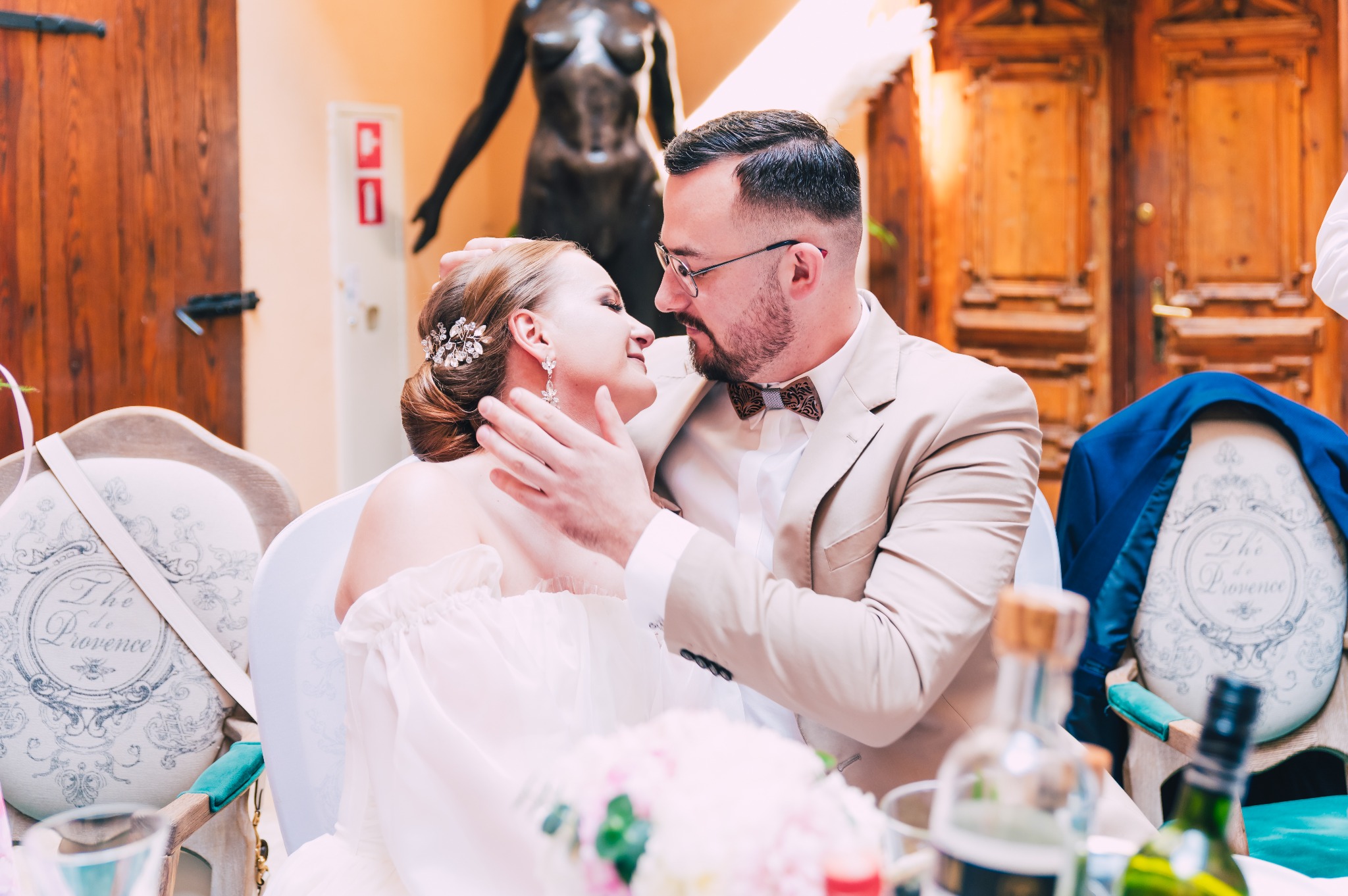 Para młoda siedzi przy stole, mężczyzna w beżowym garniturze i drewnianej muszce obejmuje kobietę w białej sukni z ozdobą we włosach, w tle drewniane drzwi i rzeźba kobiecej postaci.