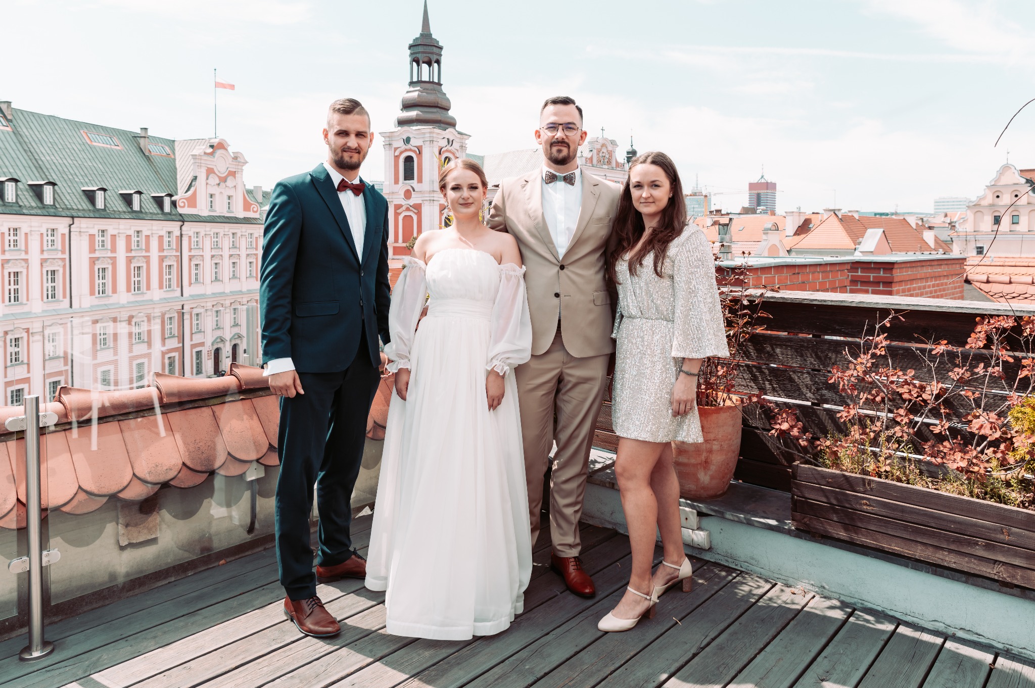 Para młoda z gośćmi na tle panoramy miasta z czerwonymi dachami i wieżą ratuszową, pozująca na drewnianym tarasie z roślinami.