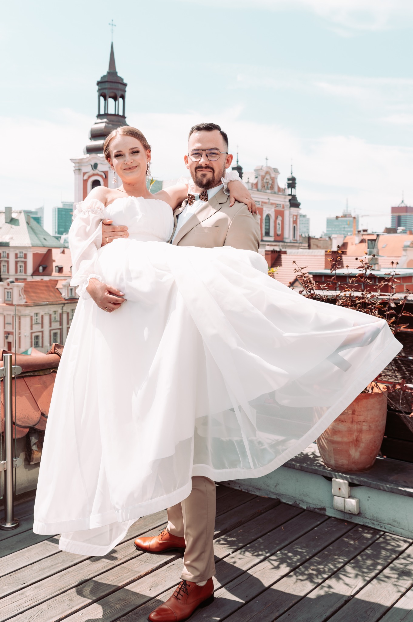 Pan młody trzyma na rękach uśmiechniętą pannę młodą w białej sukni na tle panoramy miasta z czerwonymi dachami i kościołem