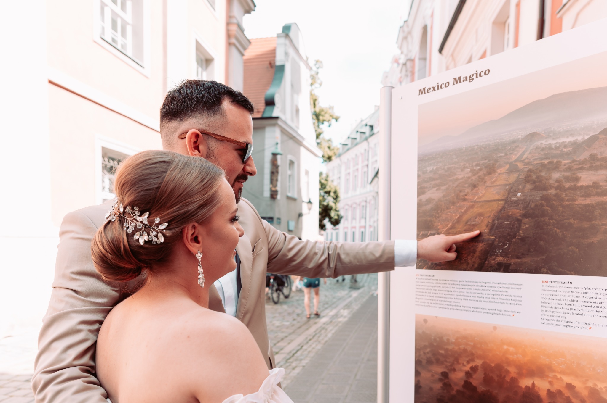 Para młoda ogląda tablicę informacyjną 'Mexico Magico' na tle miejskiej uliczki, mężczyzna wskazuje palcem na zdjęcie starożytnego miasta Teotihuacan.