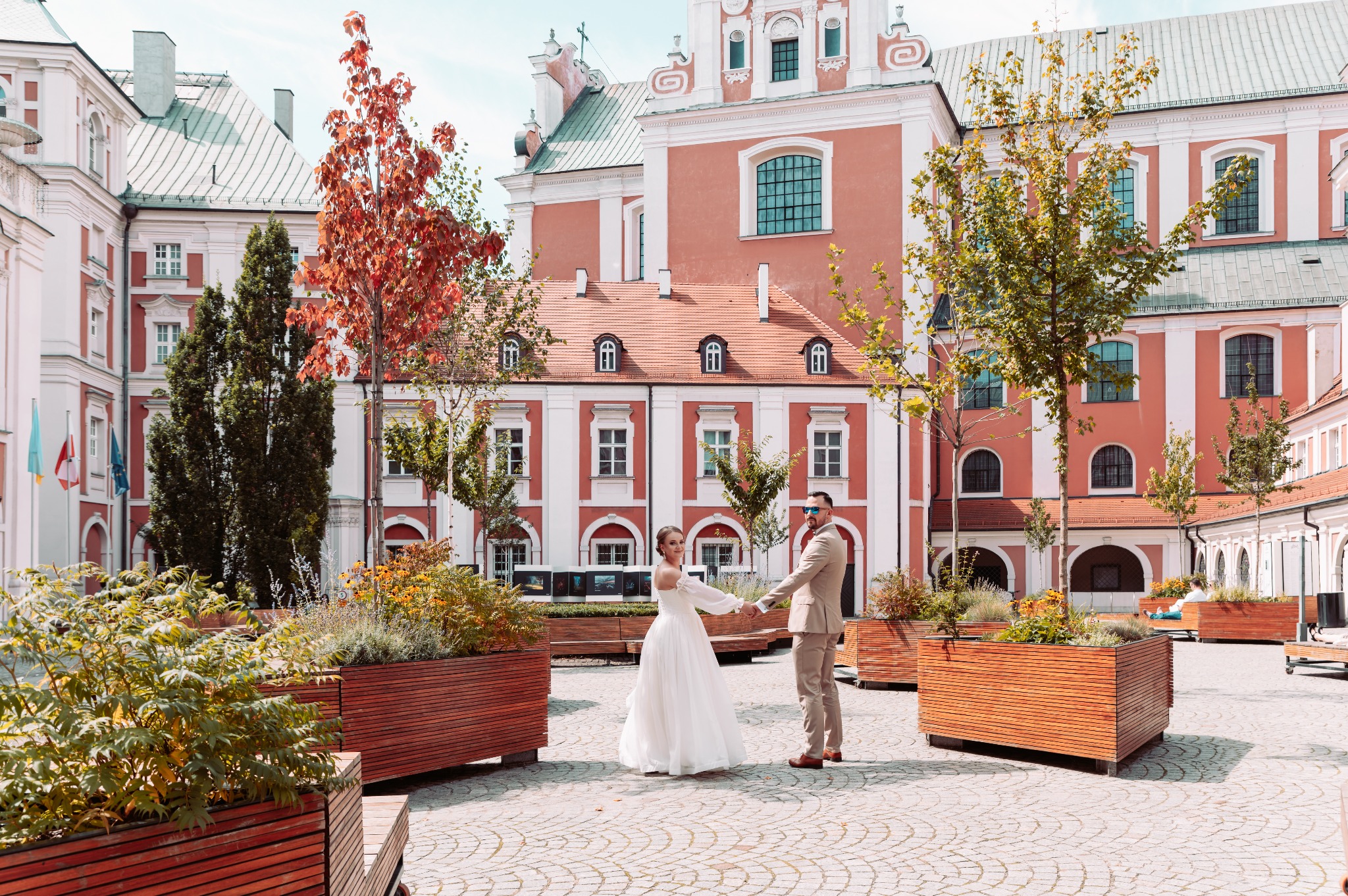 Para młoda trzymająca się za ręce na brukowanym placu, z architekturą w odcieniach czerwieni i bieli w tle oraz roślinami w drewnianych donicach