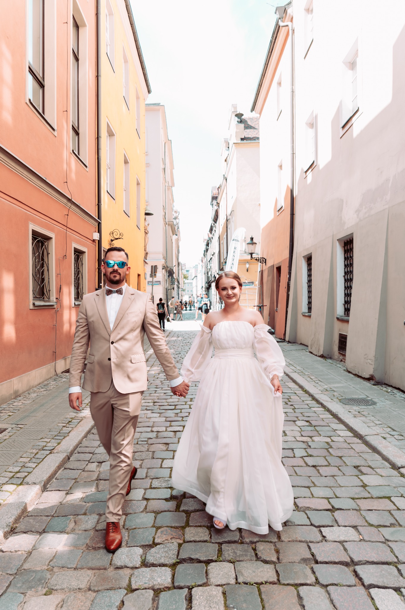 Para młoda, trzymając się za ręce, spaceruje po brukowanej uliczce w otoczeniu kamienic o pastelowych fasadach; pan młody w beżowym garniturze i okularach przeciwsłonecznych, panna młoda w zwiewnej...