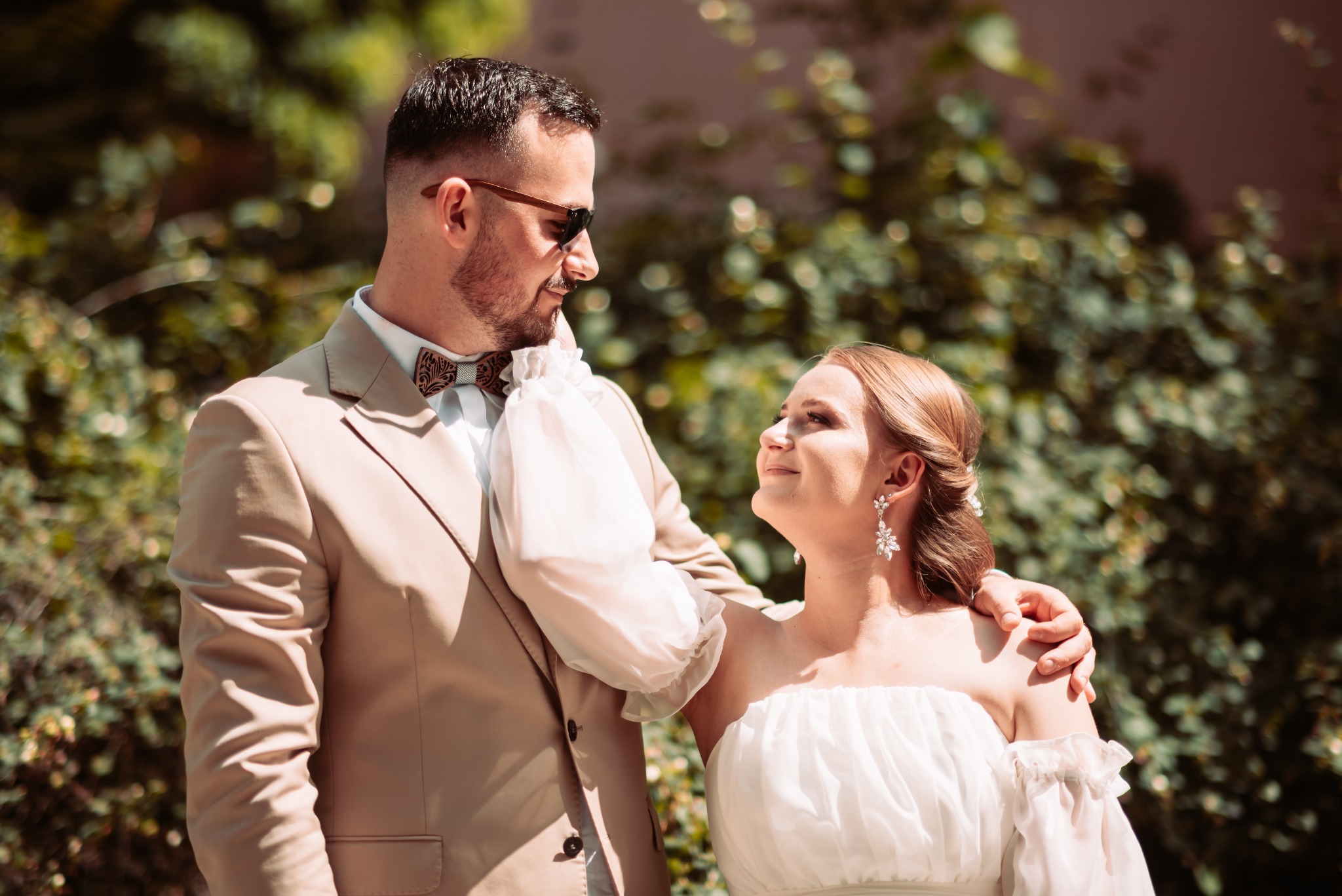 Para młoda: mężczyzna w beżowym garniturze i drewnianej muszce, kobieta w białej sukni z odkrytymi ramionami i wiszących kolczykach, oboje patrzą na siebie z uśmiechem na tle zieleni.