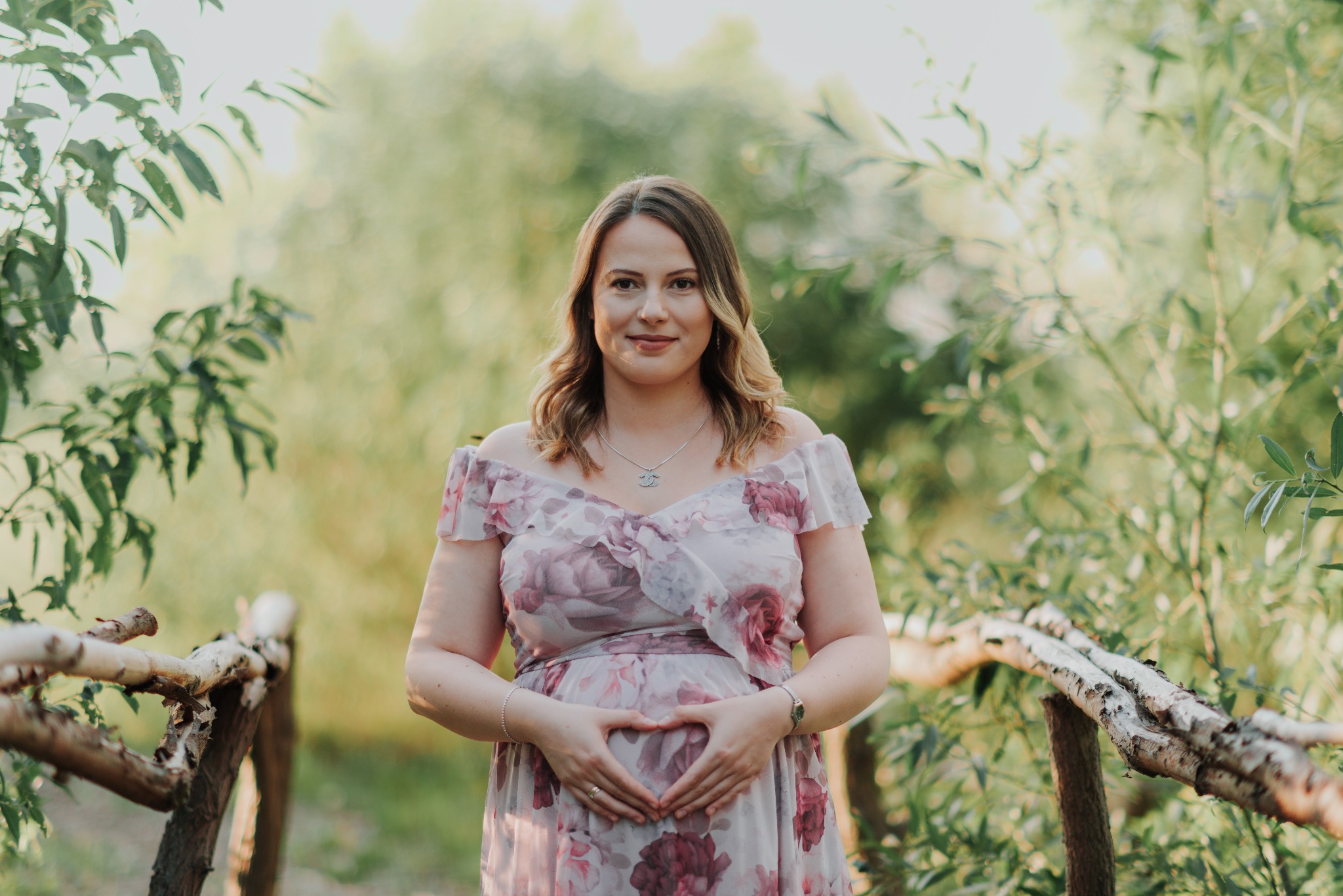 Kobieta w kwiecistej sukience ciążowej, z rękami ułożonymi w kształt serca na brzuchu, pozuje na tle zieleni i drewnianego mostku.