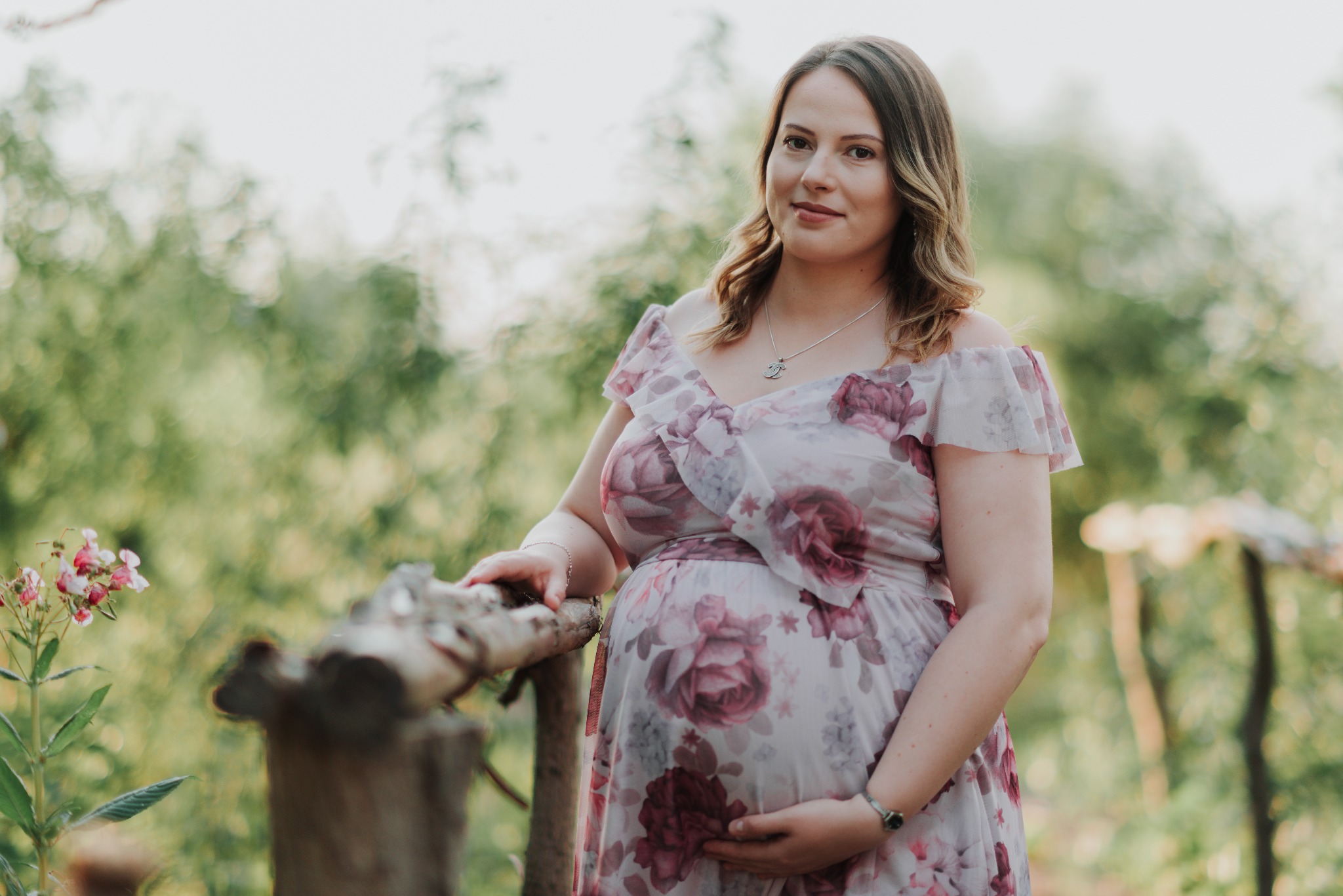 Kobieta w ciąży w kwiecistej sukience, opierająca się o drewnianą barierkę na tle rozmytej zieleni