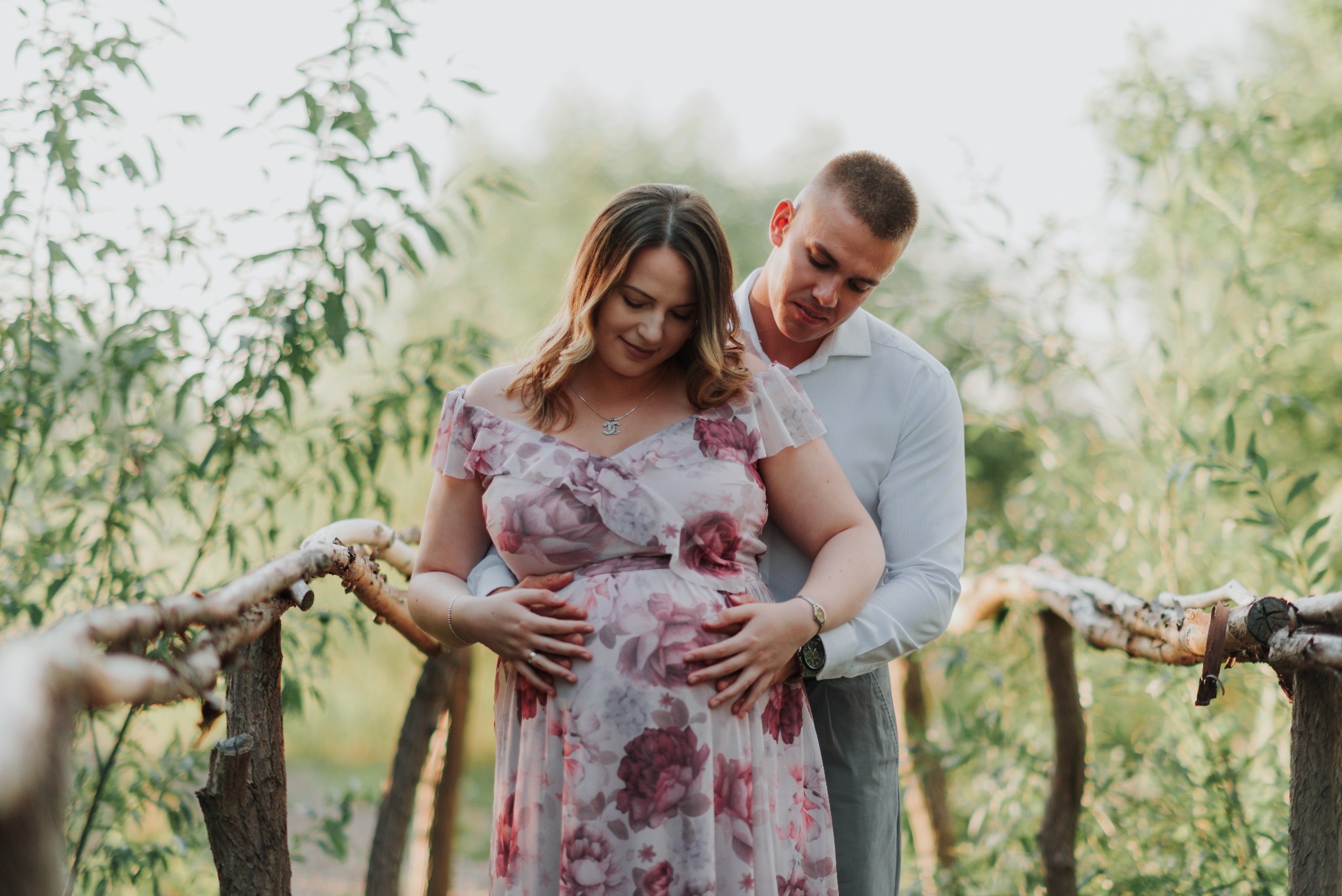 Przyszła mama w kwiecistej sukience i jej partner obejmujący jej brzuch na drewnianym mostku w otoczeniu zieleni.