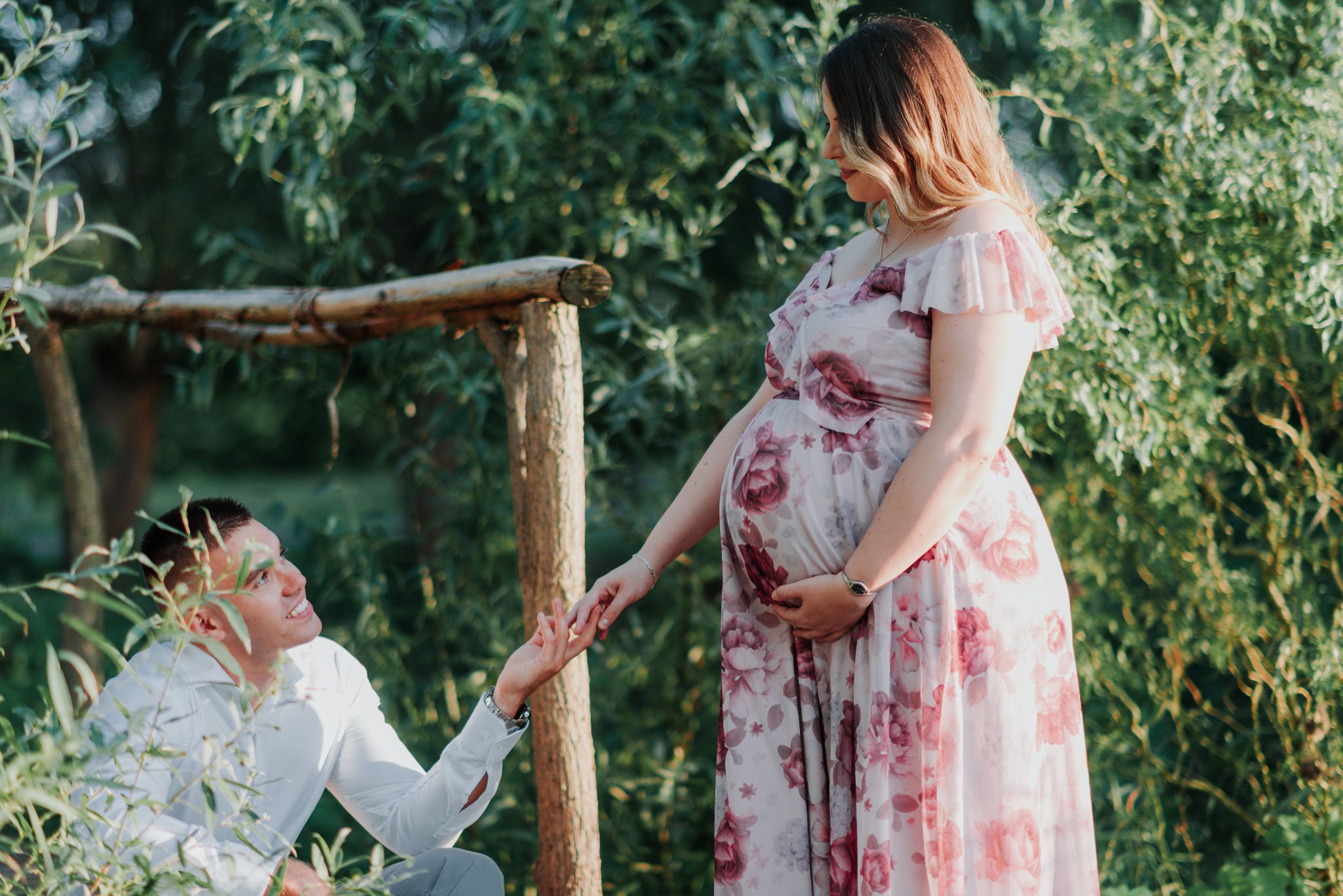 Sesja ciążowa w plenerze w Poznaniu: przyszła mama w kwiecistej sukni trzyma za rękę partnera na tle zieleni.
