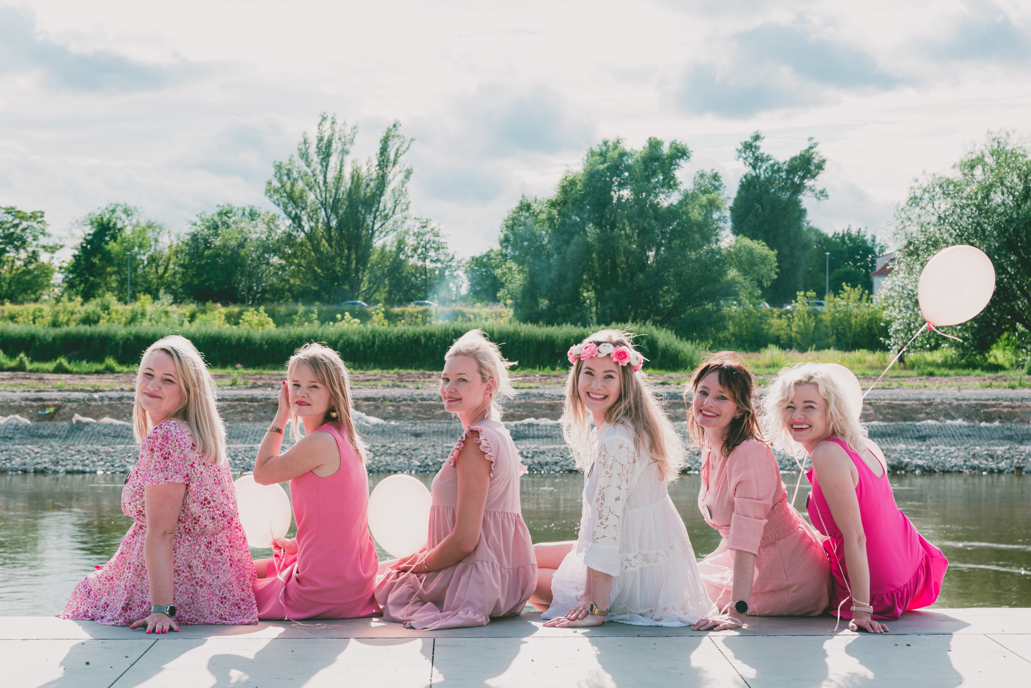 Siedzące na betonowym nabrzeżu kobiety w różowych i białych sukienkach, trzymające balony, z rzeką i zielenią w tle.