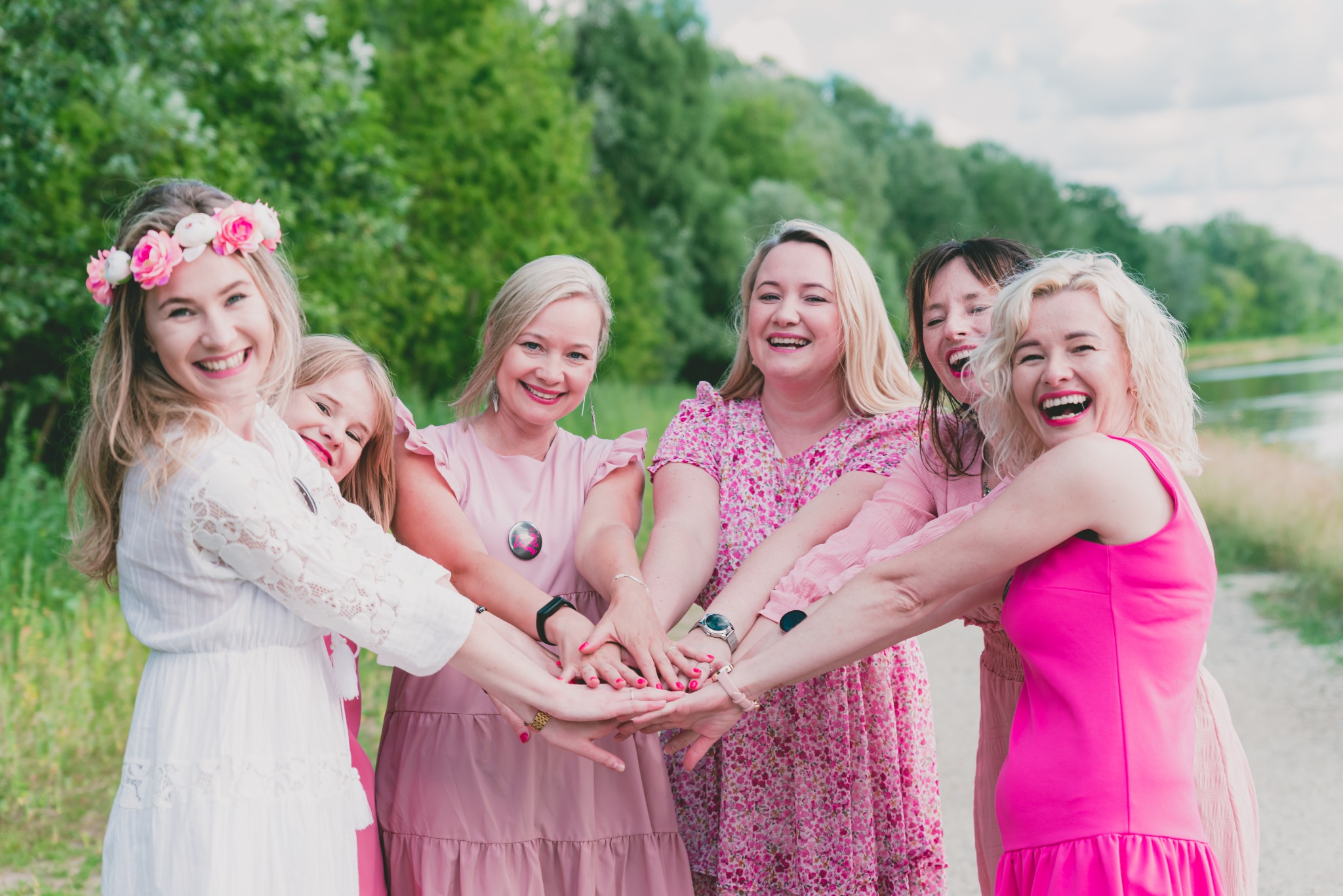 Grupa uśmiechniętych kobiet i dziewczyn w różowych sukienkach, trzymających ręce razem na tle zieleni.