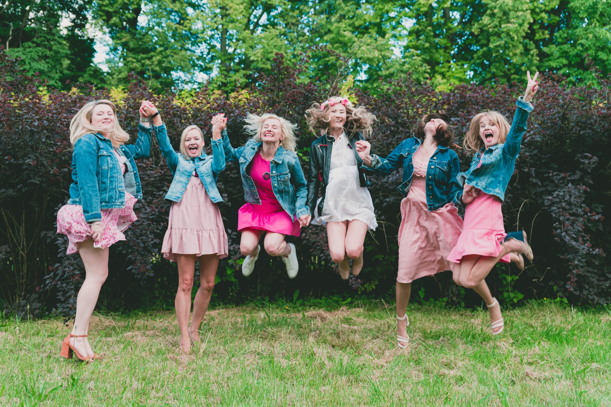 Grupa pięciu roześmianych kobiet w różowych sukienkach i jeansowych kurtkach, skaczących trzymając się za ręce na tle zieleni w plenerze w okolicach Poznania.