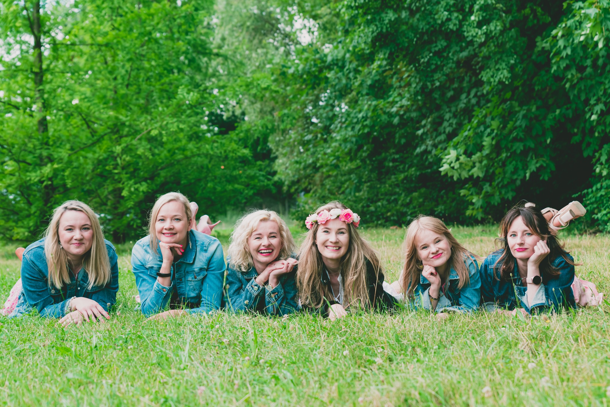 Grupa sześciu uśmiechniętych kobiet w dżinsowych kurtkach leży na trawie w parku, jedna z nich ma wianek z różowych kwiatów na głowie, w tle zielone drzewa.