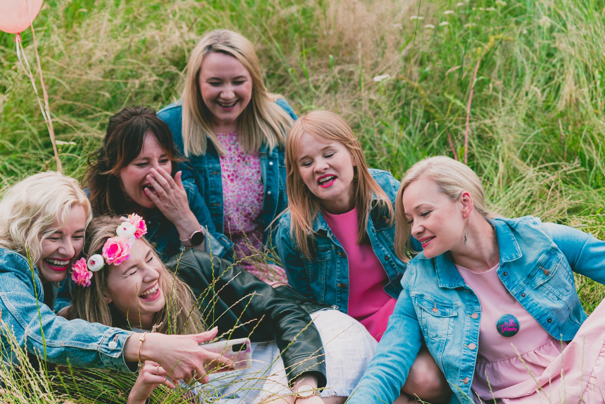 Grupa sześciu roześmianych kobiet w różowych sukienkach i jeansowych kurtkach siedzi na trawie podczas sesji zdjęciowej, jedna z nich ma wianek z różowych kwiatów, a inna przypinkę z napisem 'Bride...